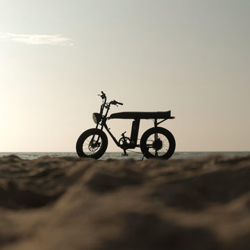 夕陽下的海邊沙丘上，一台 Unimoke 胖胎電輔車逆光停著，成為剪影