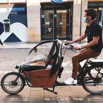 騎士在歐洲街道上騎乘前置貨箱式 Cargo Bike，車前木質貨箱裝著採買物品並罩上透明雨棚