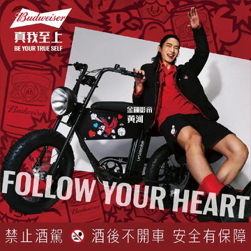 百威 × Urban Drivestyle 聯名海報：金鐘影帝黃河穿黑外套坐在一台印著愛心與老鷹插畫的黑色寬胎電輔車旁，紅色背景印著「真我至上 BE YOUR TRUE SELF」與「FOLLOW YOUR HEART」，下方是「禁止酒駕　酒後不開車　安全有保障」。