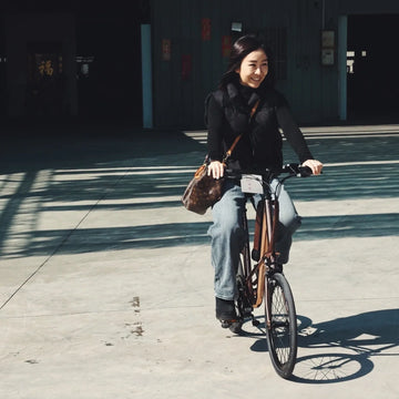 陽光下的廣場，女生騎著電輔車迎面而來，肩上背著一個包