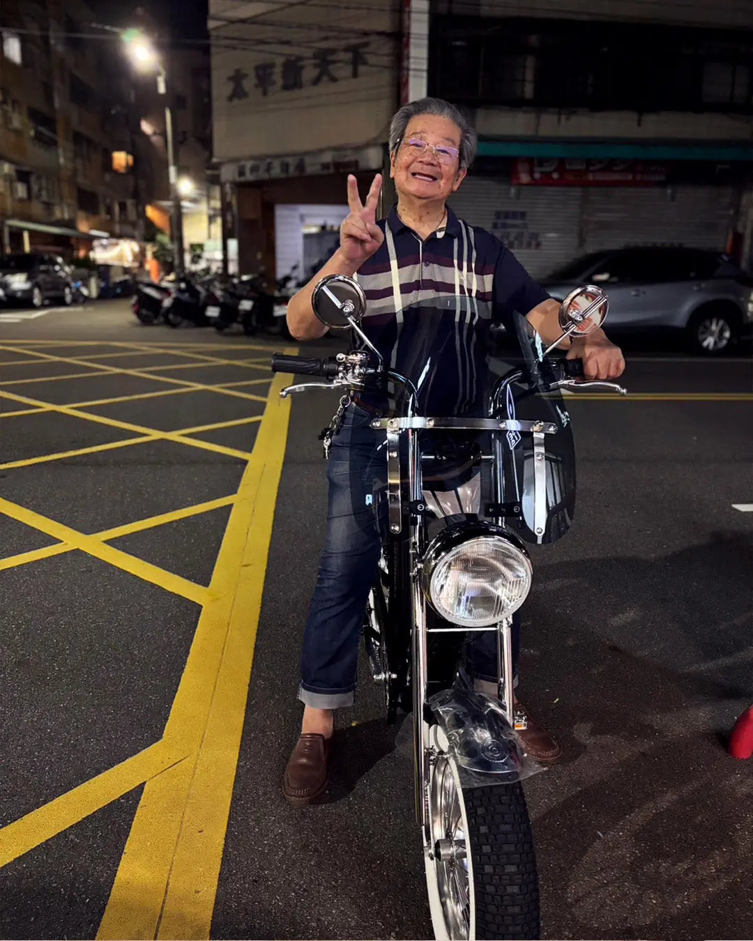 一位穿條紋 polo 衫的長者在夜間街道上跨坐於黑色電輔車，對鏡頭比出勝利手勢，背景是亮著燈的街屋與停放的機車