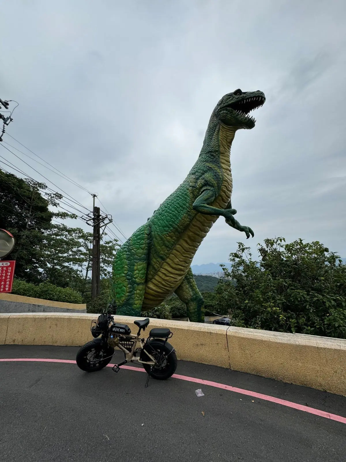 胖胎電輔車停在山路彎道的護欄前，身後是一座高聳的綠色恐龍雕像與遠方山景