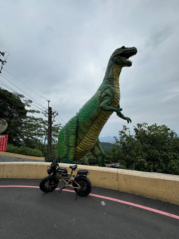 胖胎電輔車停在山路彎道的護欄前，身後是一座高聳的綠色恐龍雕像與遠方山景