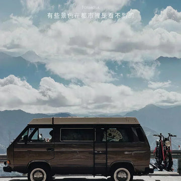 山頂公路旁停著一台深色露營車，車後架著電輔車，前方是厚厚的雲層與遠山，上方印著「有些景色在都市裡是看不到的」