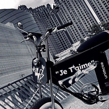 黑色小輪胖胎電輔車停在玻璃帷幕大樓前，側板噴著 Je T’aime 字樣