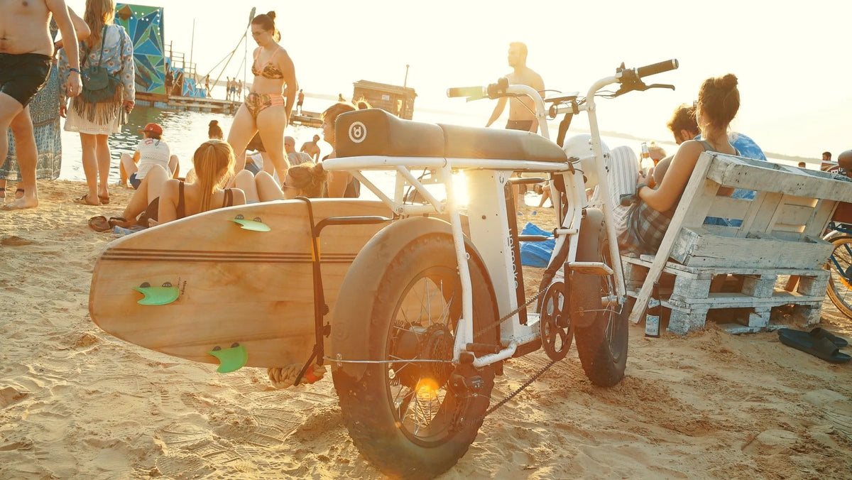 夕暮れのビーチに停めた白いE-BIKE。側面のスタンドにサーフボードを載せ、周りには砂浜でくつろぐ人たちの姿