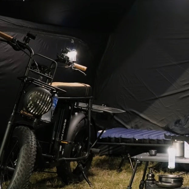 夜晚的營地，電輔車停在黑色帳篷裡，旁邊是行軍床與擺著營燈的摺疊桌