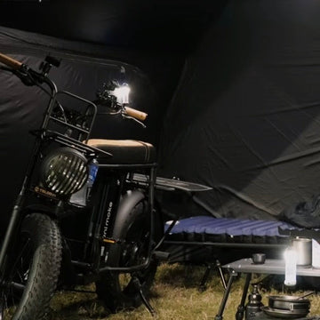 夜晚的營地，電輔車停在黑色帳篷裡，旁邊是行軍床與擺著營燈的摺疊桌