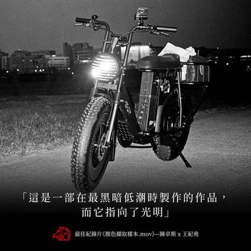 黑白夜景照片，一台胖胎電輔車停在河濱草地上亮著車頭燈，下方印著「這是一部在最黑暗低潮時製作的作品，而它指向了光明」