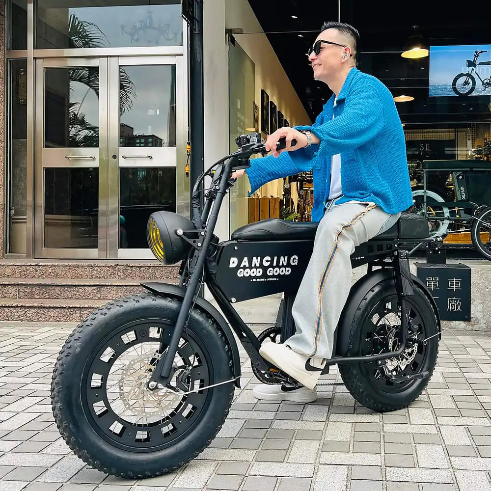 Magic 小鐘戴著墨鏡、穿藍色針織外套，坐在 SEic 單車工廠門口一台黑色寬胎 Unimoke 電輔車上。