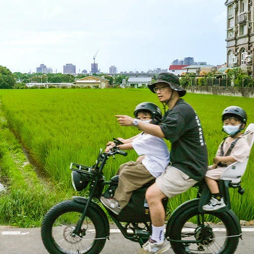 一位大人載著兩個戴安全帽的孩子騎電輔車經過稻田，遠方是城市天際線