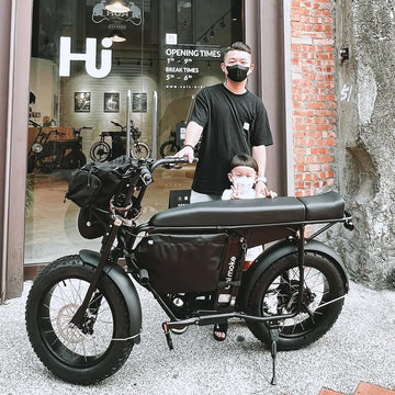 父親牽著裝有前置行李包與長坐墊的黑色電輔車，孩子坐在車上，停在紅磚外牆的店家門口