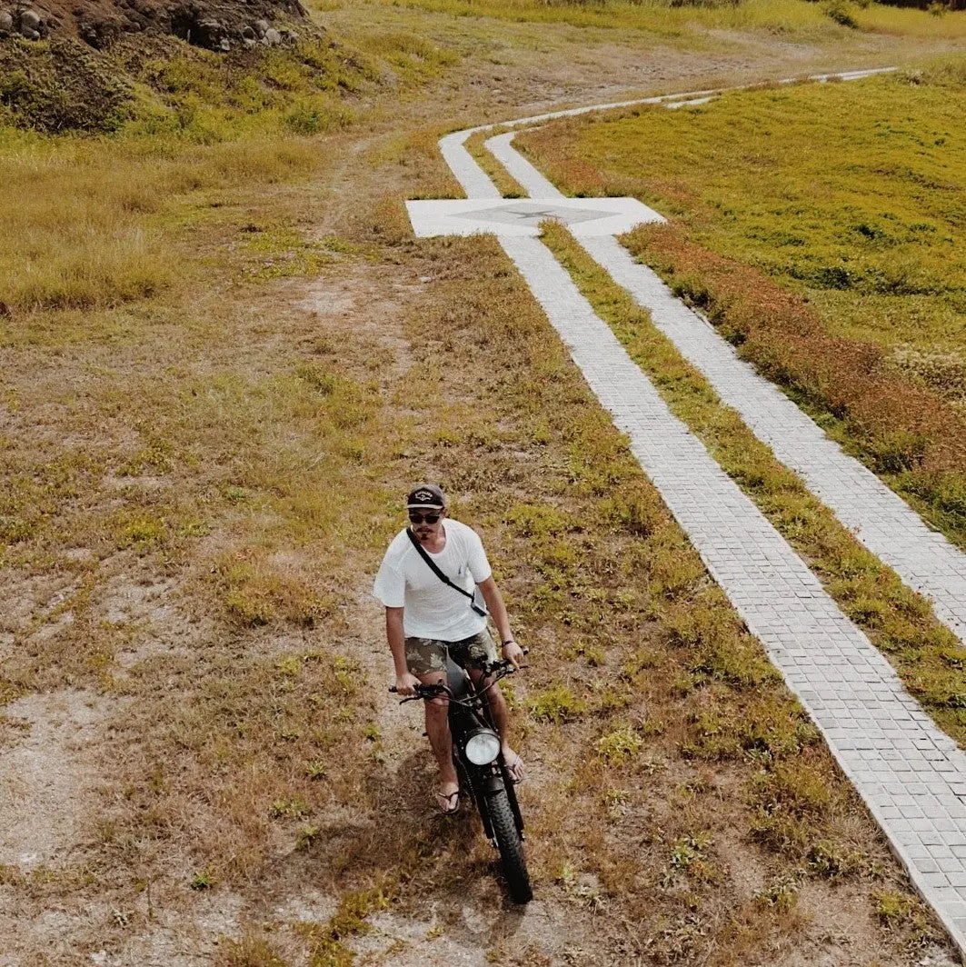 空拍視角，一位騎士騎著胖胎電輔車行經草地間的白色步道，兩側是枯黃的草坡