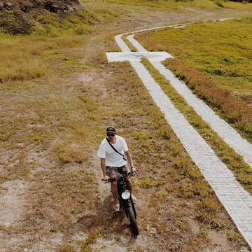 空拍視角，一位騎士騎著胖胎電輔車行經草地間的白色步道，兩側是枯黃的草坡
