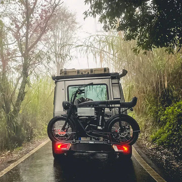 越野露營車停在雨後的山間林道上，車尾架著一台黑色胖胎電輔車，兩側是芒草與霧氣