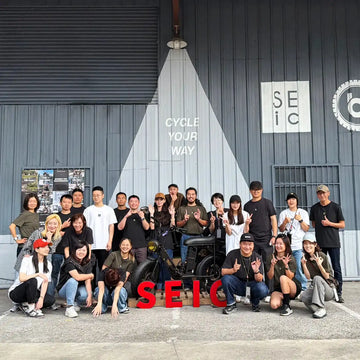 SEic 全體員工在工廠大門前合照，牆上寫著 CYCLE YOUR WAY，地上擺著紅色 SEIC 立體字
