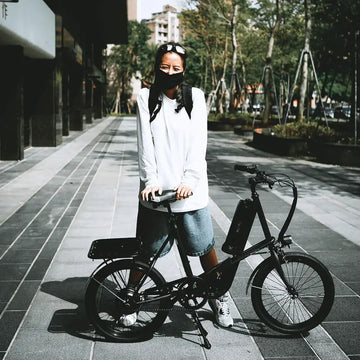一位戴口罩、穿白色長版上衣與丹寧短褲的女生，牽著一台黑色 miniu 折疊電輔車站在人行道上，背景是行道樹與大樓。