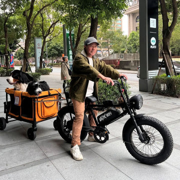 戴鴨舌帽的男士跨坐在 UDX 電輔車上，旁邊的橘色寵物拖車裡坐著一隻伯恩山犬和一隻黑白長毛狗，背景是街道與行道樹