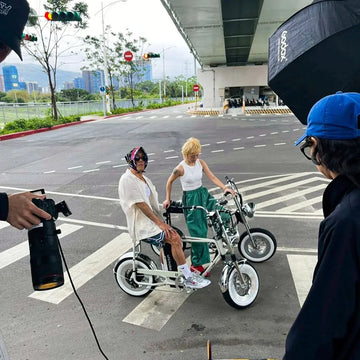 拍攝現場側拍，兩位模特兒站在路口的斑馬線上與兩台電輔車合影，前景是攝影師的相機與燈具