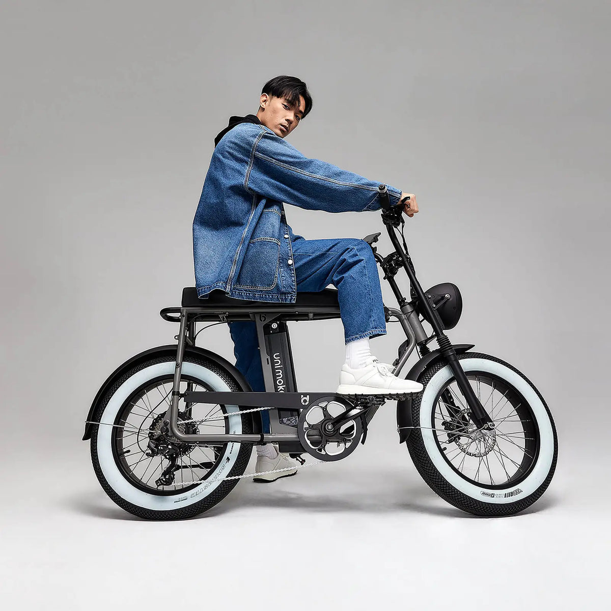 一名男子身著牛仔褲和牛仔外套，騎乘白邊胎的電輔車 Unimoke復古銀，時尚且帥氣
